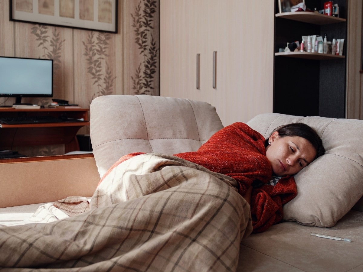 Woman resting under blankets while keeping an eye on a thermometer beside her, representing common cold and flu symptoms.