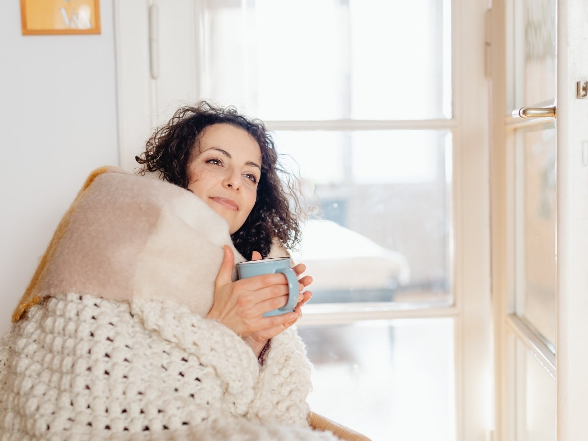 Woman wrapped in a blanket holding a warm mug, showing a calm and comfortable recovery environment during winter.